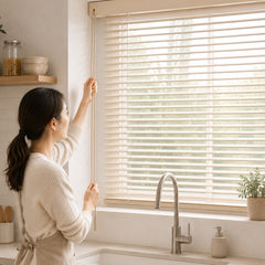 A woman in a cream ribbed sweater and matching apron adjusts a beige roller blind system on a window above a kitchen sink.